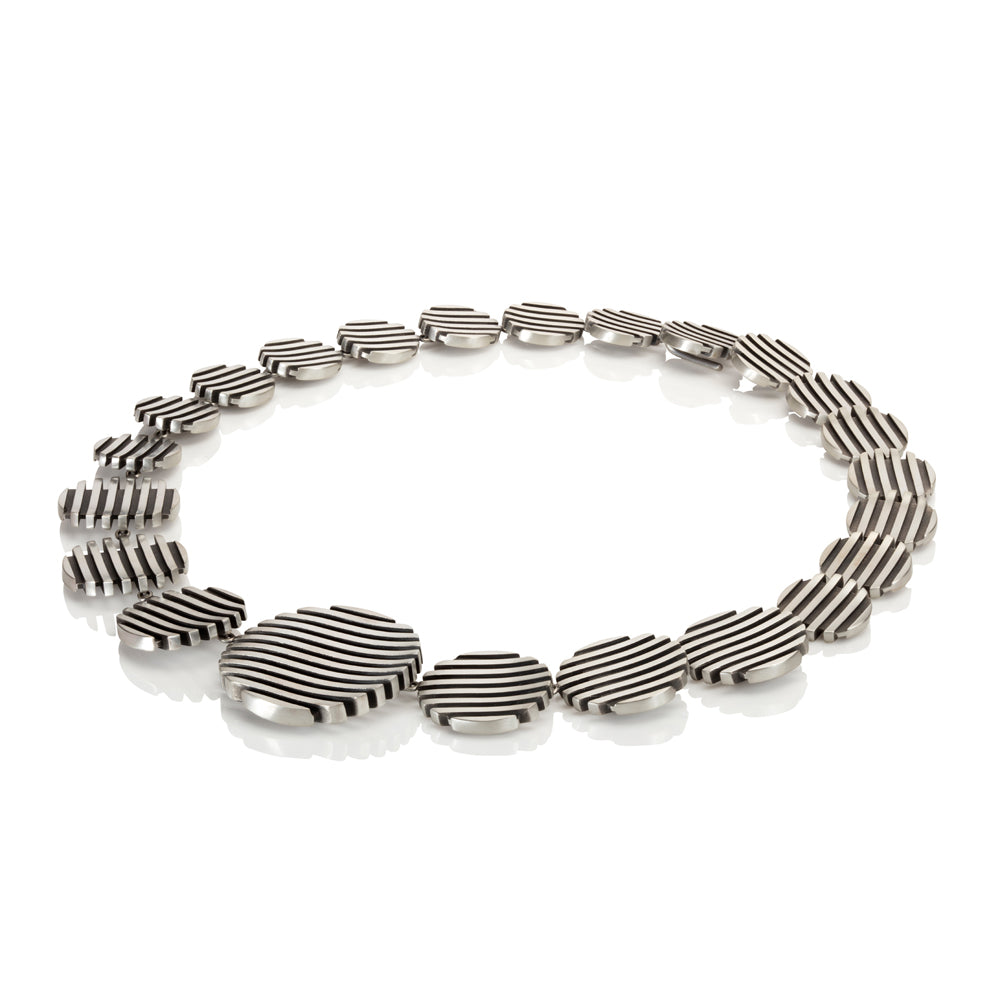 The escalator neckpiece shows geometric abstraction through its precisely offset tapering disc construction, the gently undulating surfaces create a dynamic interplay of light, while the oxidised recesses articulate the linear deconstructed form. Escalator neckpiece by Sarah Herriot.