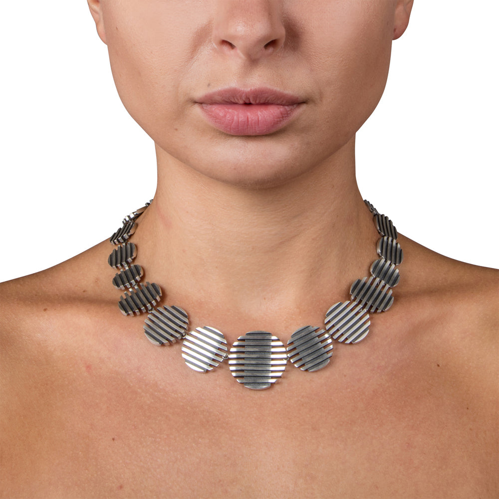 The escalator neckpiece shows geometric abstraction through its precisely offset tapering disc construction, the gently undulating surfaces create a dynamic interplay of light, while the oxidised recesses articulate the linear deconstructed form. Escalator neckpiece by Sarah Herriot.