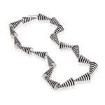 This striking neckpiece in silver showcases an inventive architectural approach to contemporary jewellery design, composed of linked pyramid forms, the piece uses linear deconstruction to create visual rhythm and movement. Oxidised recesses accentuate and amplifying the jazzy aesthetic, a bold statement piece for those seeking unconventional jewellery that challenges traditional forms whilst maintaining refined craftsmanship. Pyramid neckpiece by Sarah Herriot.