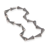 This striking neckpiece in silver showcases an inventive architectural approach to contemporary jewellery design, composed of linked pyramid forms, the piece uses linear deconstruction to create visual rhythm and movement. Oxidised recesses accentuate and amplifying the jazzy aesthetic, a bold statement piece for those seeking unconventional jewellery that challenges traditional forms whilst maintaining refined craftsmanship. Pyramid neckpiece by Sarah Herriot.